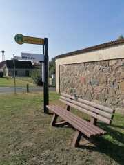 braune Parkbank mit Beschilderung Mitfahrerbank grüne Schrift auf gelben Hintergrund, darunter Schild Richtung Blindow aufgeklappt, im Hintergrund eine Feldsteinmauer