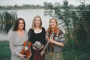 Drei Musikerinnenstehen mit Geige, Ziehharmonika und Flöte vor einem See