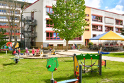 Spielplatz der Kita Kinderland, Spielgeräte, Sandkasten, Überdachter Sitzbereich, Gebäude mit Außenrollos und Feuertreppe 