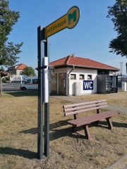 braune Parkbank mit Beschilderung Mitfahrerbank grüne Schrift auf gelben Hintergrund, im Hintergrund ein WC Häuschen mit Snackautomaten