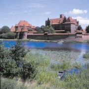 Blick auf eine alte Burg, im Vordergrund Wiese und Wasser