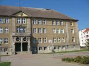 4 stöckiges Gebäude mit Verzierungen an der Betonfassade und weißen Sprossenfenstern, Treppe zu 2 großen Eingangstüren vor diesen ist ein Balkon für die 3 Etage gebaut, dunkles Spitzdach mit Antenne, Hof gepflastert, ein Basketballkorb auf dem Hof