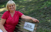 Frau Meinke sitzt auf einer Parkbank auf der einer silbernen Plattdeutsch-Tafel mit Verzieherungen am Rand und der Aufschrift: Fleuten am morgen, jifft Kummer un Sorgen.