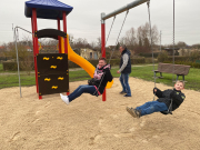 Sandplatz mit Doppelschaukel auf der eine Frau und ein Junge schaukeln, Parkbank, ein Rutschenturm mit Kletterwand und Rutschstange