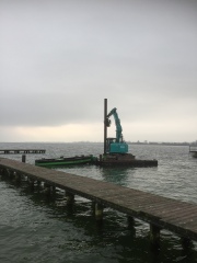 mit einer schwimmenden Pfahlramme werden die Pfähle gesetzt, im Vordergrund ist der alte Steg zusehen, ein grün, schwarzes Boot liegt zwischen Steg und der schwimmenden Plattform mit dem Spezialbagger 