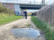 Herr Stübs mit dem Fahrrad auf dem Sandweg der Magarinefabrik mit großer Pfütze im Vordergrund